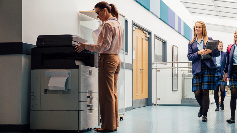 School teacher using a photocopier, children in corridor, smiling